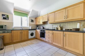 A kitchen with wooden cabinets, a washing machine, a microwave, and a window with a green blind.