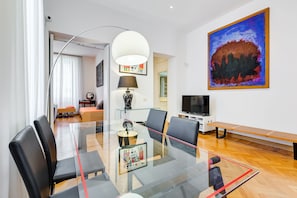 A modern living room with a glass dining table, black chairs, a sofa, a TV, and a large abstract painting.