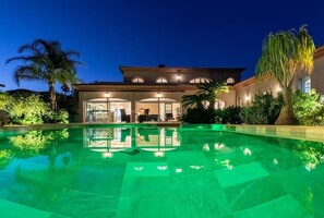 Una piscina amplia y bien iluminada con un reflejo claro, rodeada de exuberante vegetación y un edificio grande de varios pisos con ventanales arqueados y techo de tejas.