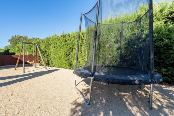 Un trampolín con red de seguridad, sobre una superficie de grava, rodeado de follaje y un columpio al fondo.