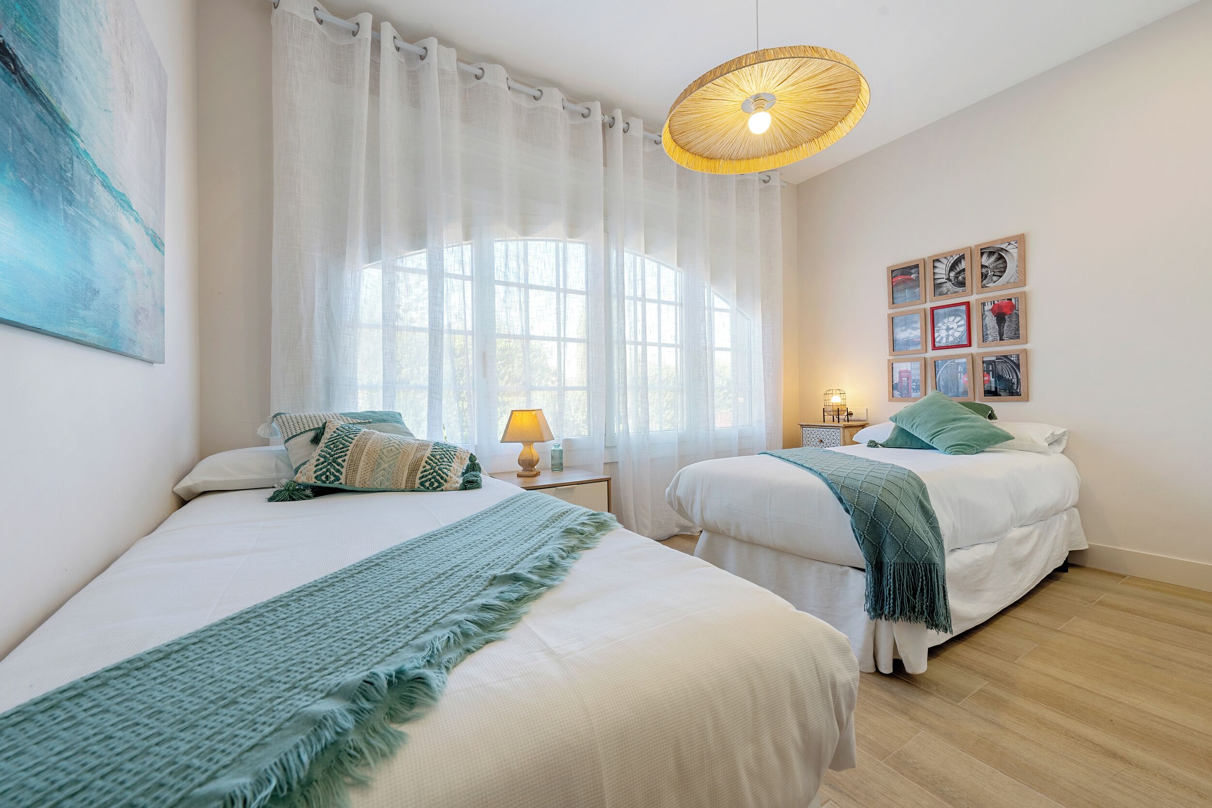 Een slaapkamer met twee bedden, een raam met dunne gordijnen, een wandlamp en kunstwerken in lijsten.