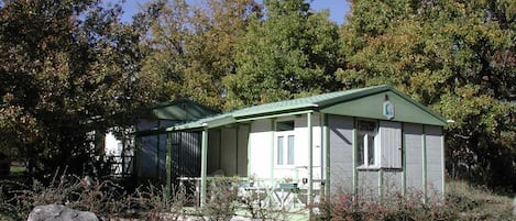 A small, single-story cabin with a green roof and white walls, surrounded by trees and rocks.
