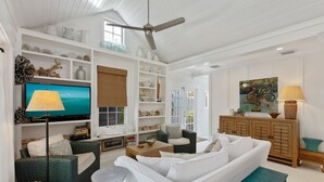 A spacious living room with a white sofa, green wicker chairs, a wooden coffee table, and a television on a stand. Shelves with decorative items line the walls. - QUEEN OF SOL ~ 3 Bed, 2 Bath in Peaceful Neighborhood with Dip Pool! (Key West)
