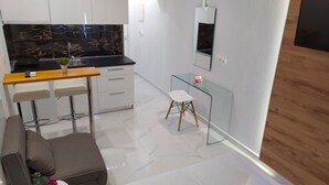 Una cocina moderna con protector contra salpicaduras de mármol negro, armarios blancos y encimera de madera. Hay un pequeño comedor con una mesa de vidrio y una silla blanca.