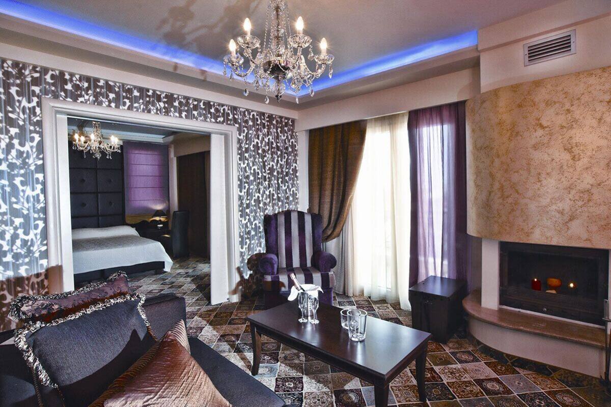 A modern hotel room with a large bed, a seating area with a sofa and armchairs, a fireplace, and a patterned floor.