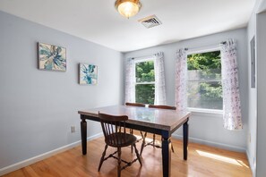 A dining area with a wooden table and chairs, two windows with floral curtains, and abstract paintings on the wall.