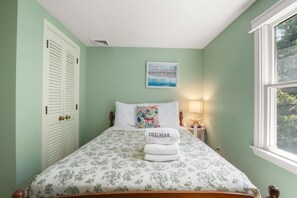 A neatly made bed with a floral bedspread and a DELMAR vacation rentals sign, a window with a view of greenery, and a wall-mounted painting. - Mins to Nauset Beach & Walkable to Town! (Orleans)