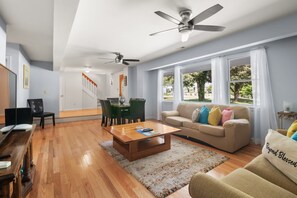A spacious living room with a wooden coffee table, a beige sofa with colorful cushions, a dining area with a wooden table and chairs, and a ceiling fan.