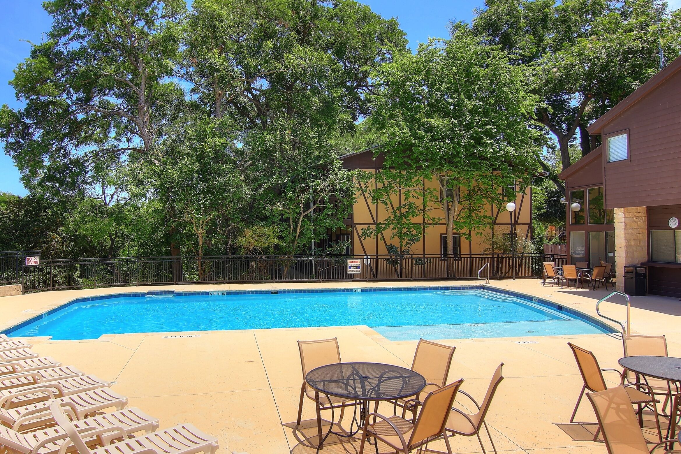 A swimming pool with lounge chairs and tables, surrounded by trees and a building with a balcony.