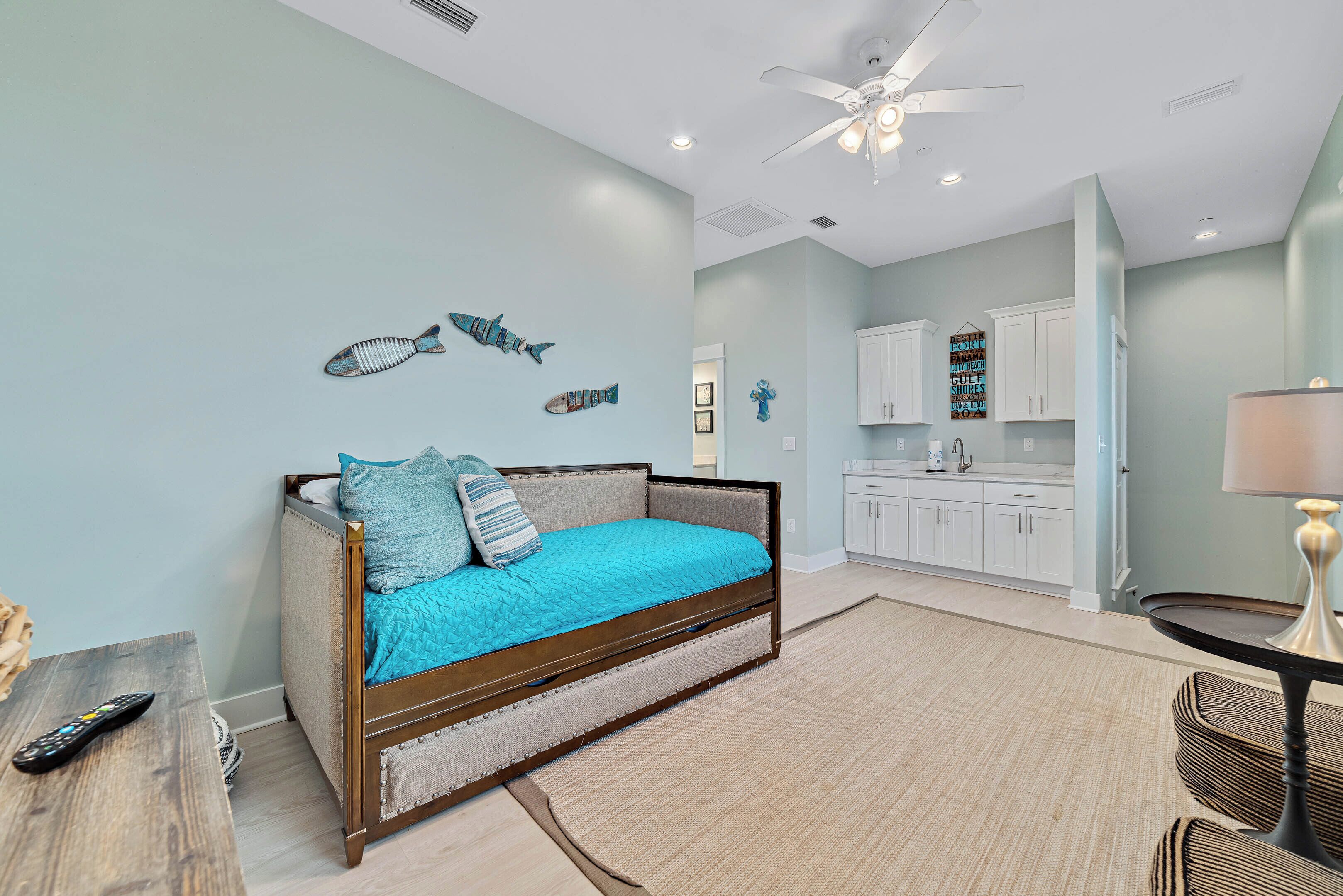 A bedroom with a bed, a ceiling fan, and a kitchenette in the background.