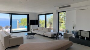 A modern living room with a sofa, ottoman, and coffee table, large windows with a view of the sea, and a flat-screen TV mounted on the wall. - Luxury villa Colomba in Brela, seafront (Brela)