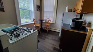 A compact kitchen with a gas stove, a refrigerator, and wooden cabinets. There are two windows with blinds, a small table, and two chairs. - Located in the Historic downtown district, cozy studio, wifi. (Auburn)