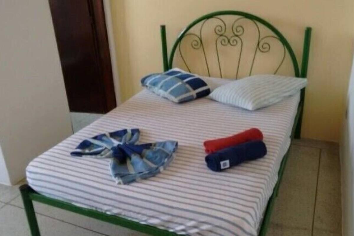 Una cama individual con colchón a rayas, cabecera de metal verde y dos almohadas.
