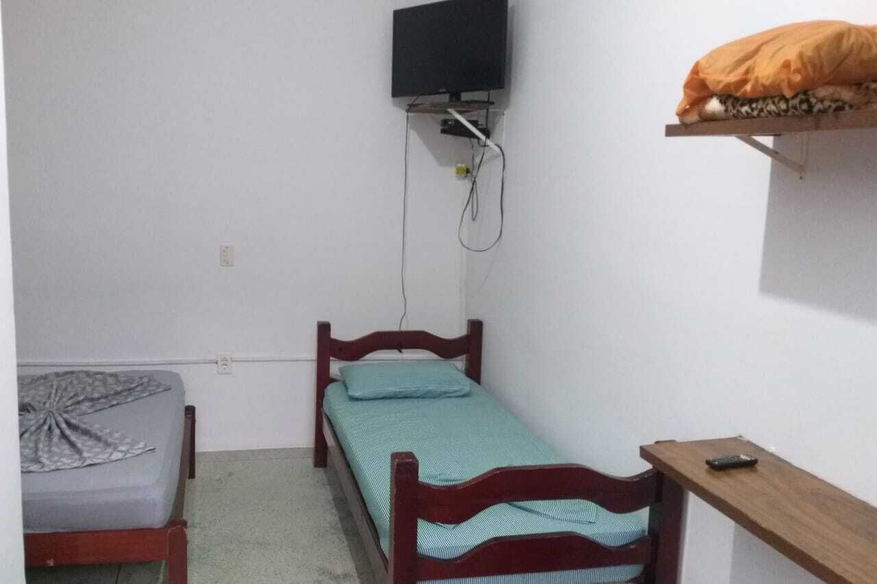 A small, sparsely furnished room with a single bed, a television mounted on the wall, and a shelf with a bag.