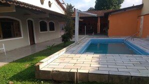 A swimming pool with a tiled deck, surrounded by a grassy area and a building with a tiled roof and arched windows. - Pousada Nova Opção (Bertioga)