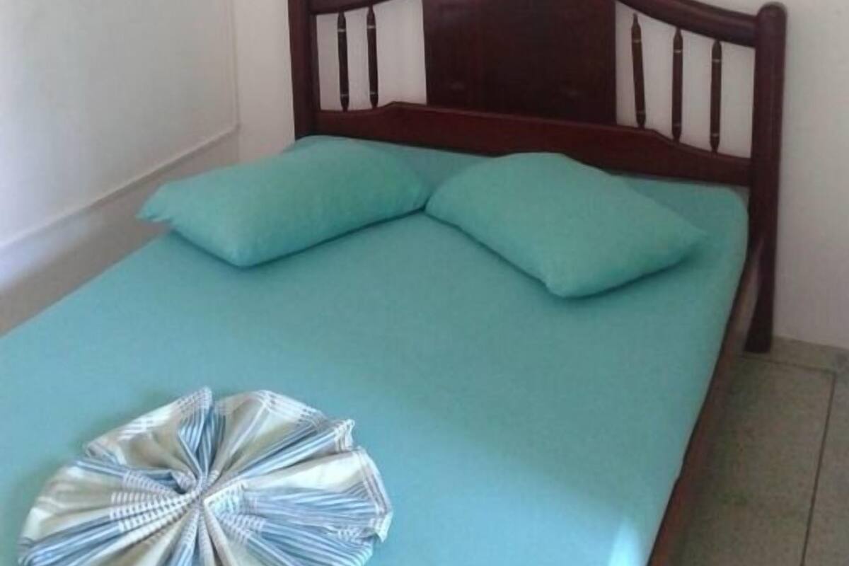 Una cama individual con cabecera de madera, una colcha color aguamarina y dos almohadas color aguamarina. Un abanico decorativo cuelga sobre la cama, y hay un cuadro enmarcado de un atardecer en la pared.