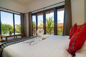 A hotel room with a large bed, a wooden nightstand, and a view of palm trees and a red-roofed building.