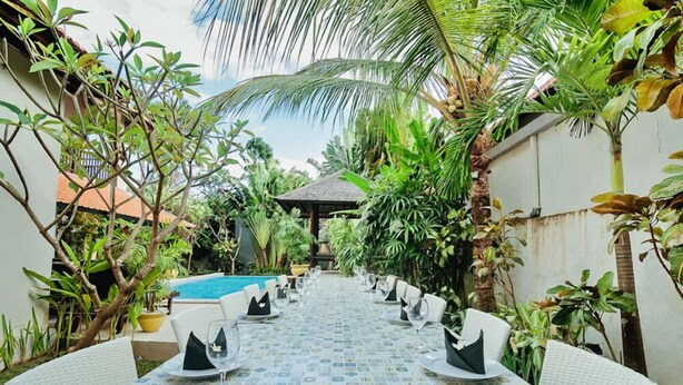 A dining area with a long table set for a meal, surrounded by lush greenery and a pool in the background. - Experience Mawar Estate, a 7-bedroom private villa estate in Seminyak. Accommodates up to 14 guest, includes daily breakfast, a full-time private chef, airport transfers, complimentary massages, housekeeping, and a dedicated driver. (Seminyak)