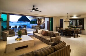 A spacious living area with a large sofa set, a coffee table, and a dining table. There is a view of the ocean through large glass doors.
