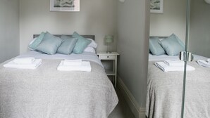 Et hotelværelse med to senge, hver med et gråt dynedække og turkise puder, et sengebord med en lampe og et blomsterbillede på væggen.