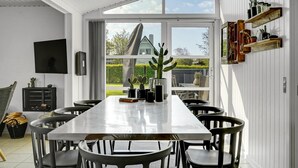 Ein Essbereich mit einem langen Tisch, Stühlen, einem Fernseher und einem Fenster mit Blick auf ein Haus und einen Garten.