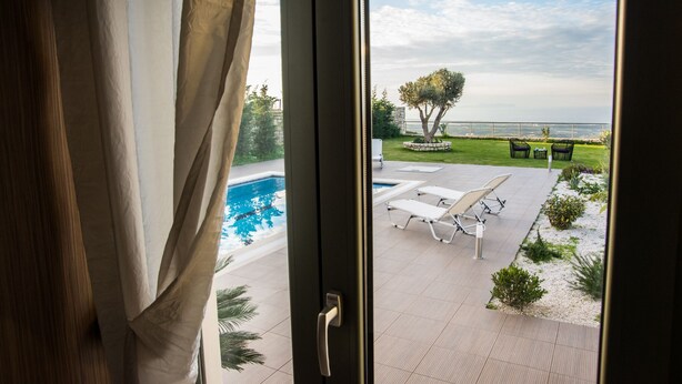 A view through a window of a pool area with lounge chairs and a tree. - Modern Villa with Panoramic Sea Views (8JQW+PXC Arkadi, Greece)