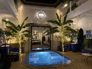 A pool area with a tiled floor, surrounded by large potted plants and blue chairs.