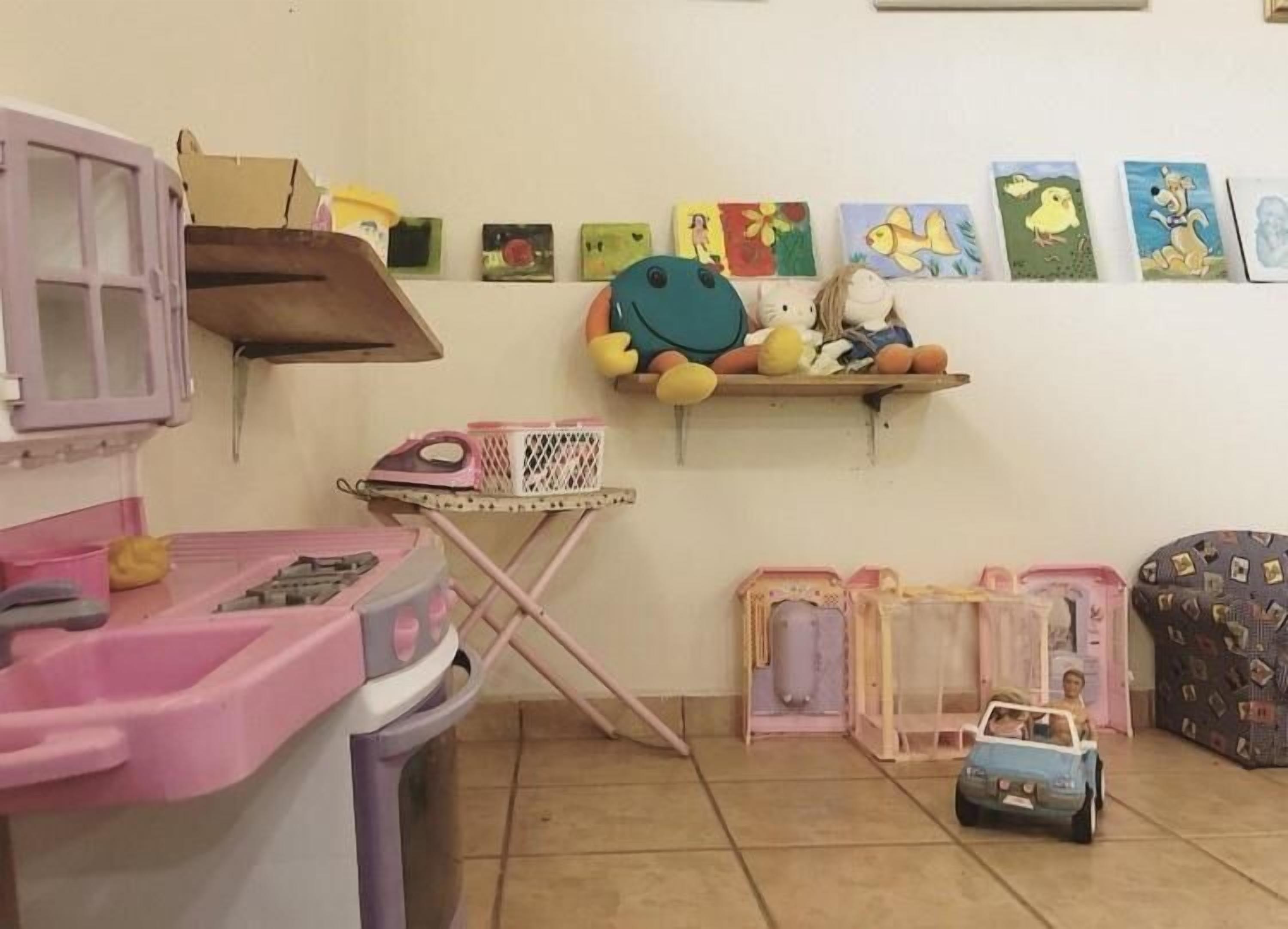 Un cuarto de juegos infantil con una cocina de juguete rosa, una tabla de planchar pequeña y un estante con dibujos hechos por niños.