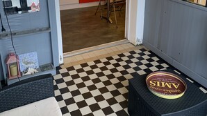 A room with a checkered floor, a small table, a chair, a lamp, a wicker chair, a small table with a cake, and a kitchen visible through an open door.