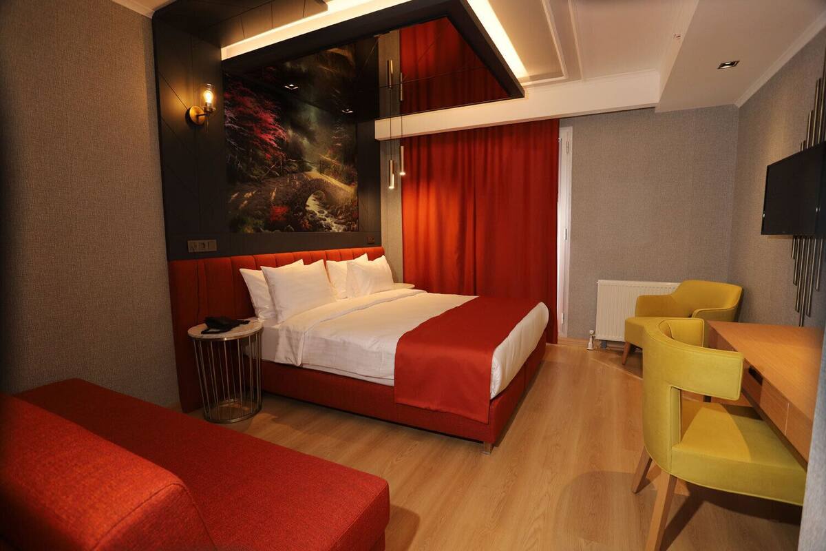 A hotel room with a bed, a red sofa, a yellow chair, a small table, and a painting on the wall.