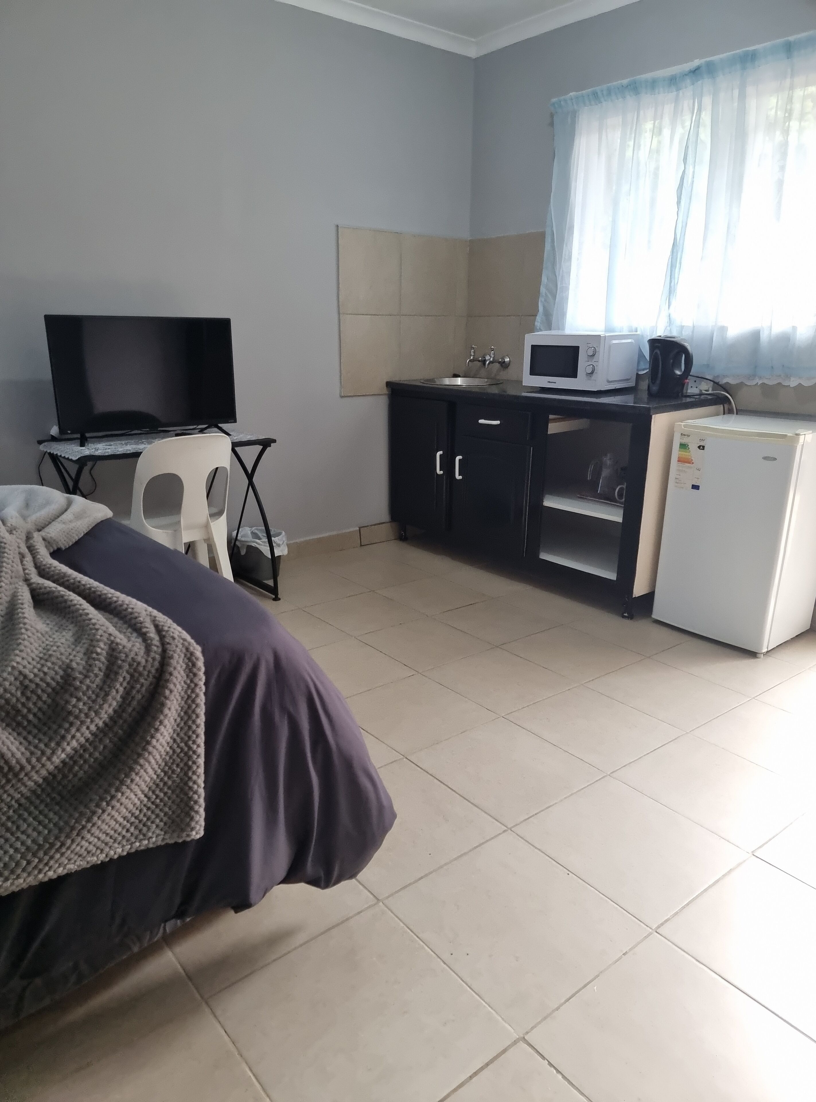 A compact hotel room with a bed, a desk with a computer and a TV, a kitchenette with a microwave and a fridge, and a window with sheer curtains.