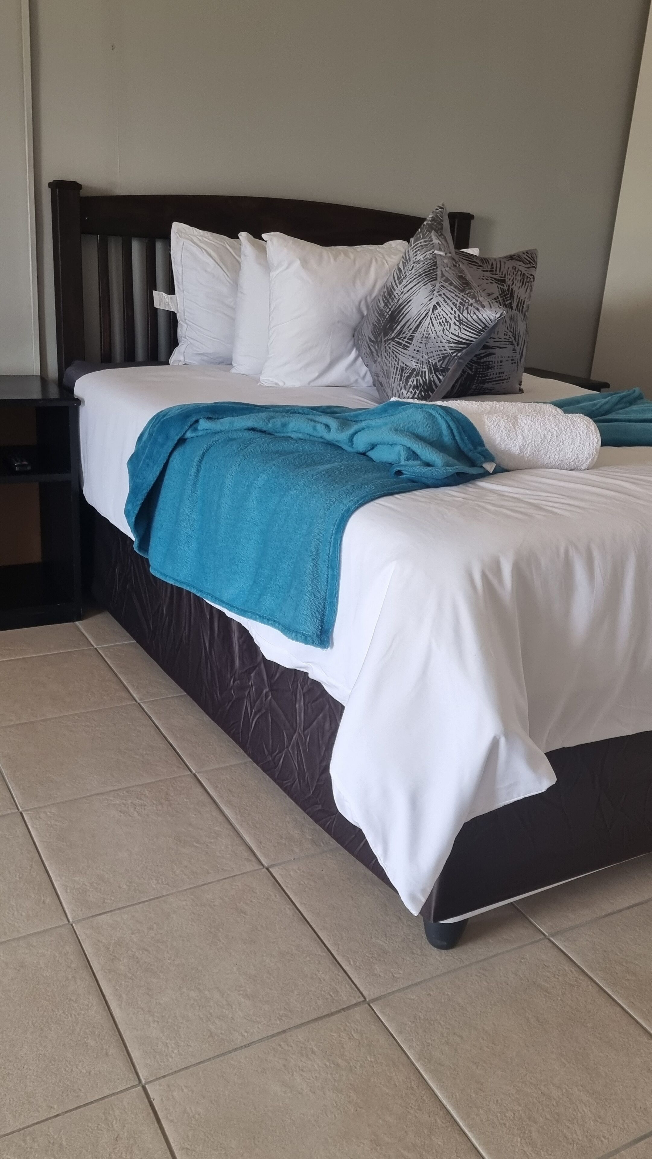 A neatly made bed with white bedding and a teal throw blanket, flanked by a dark wooden headboard and a nightstand with a lamp.