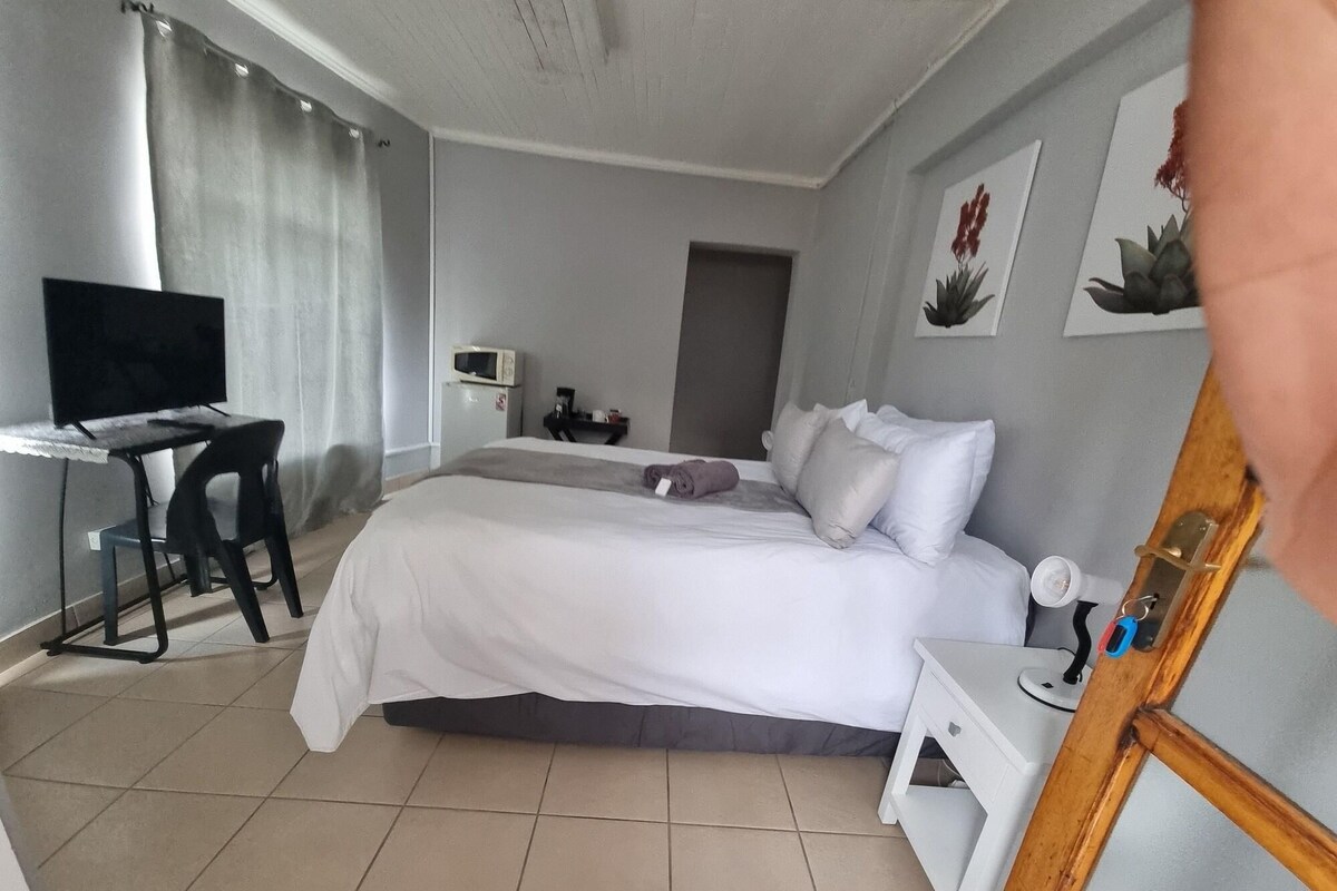 Quarto de hotel com cama, escrivaninha, televisão, cadeira, mesa pequena e porta.