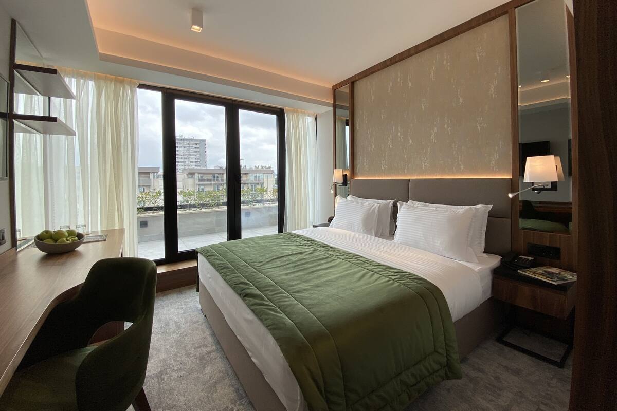 A hotel room with a large bed, a desk with a chair, a mirror, and a view of the city through large windows.