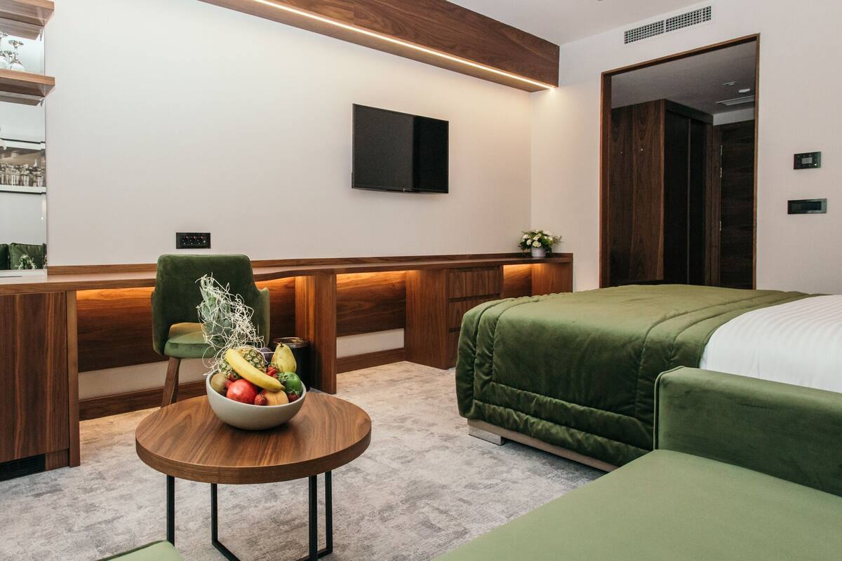 A modern hotel room with a wooden desk, a green sofa, a bed with a green cover, and a fruit bowl on a round table.