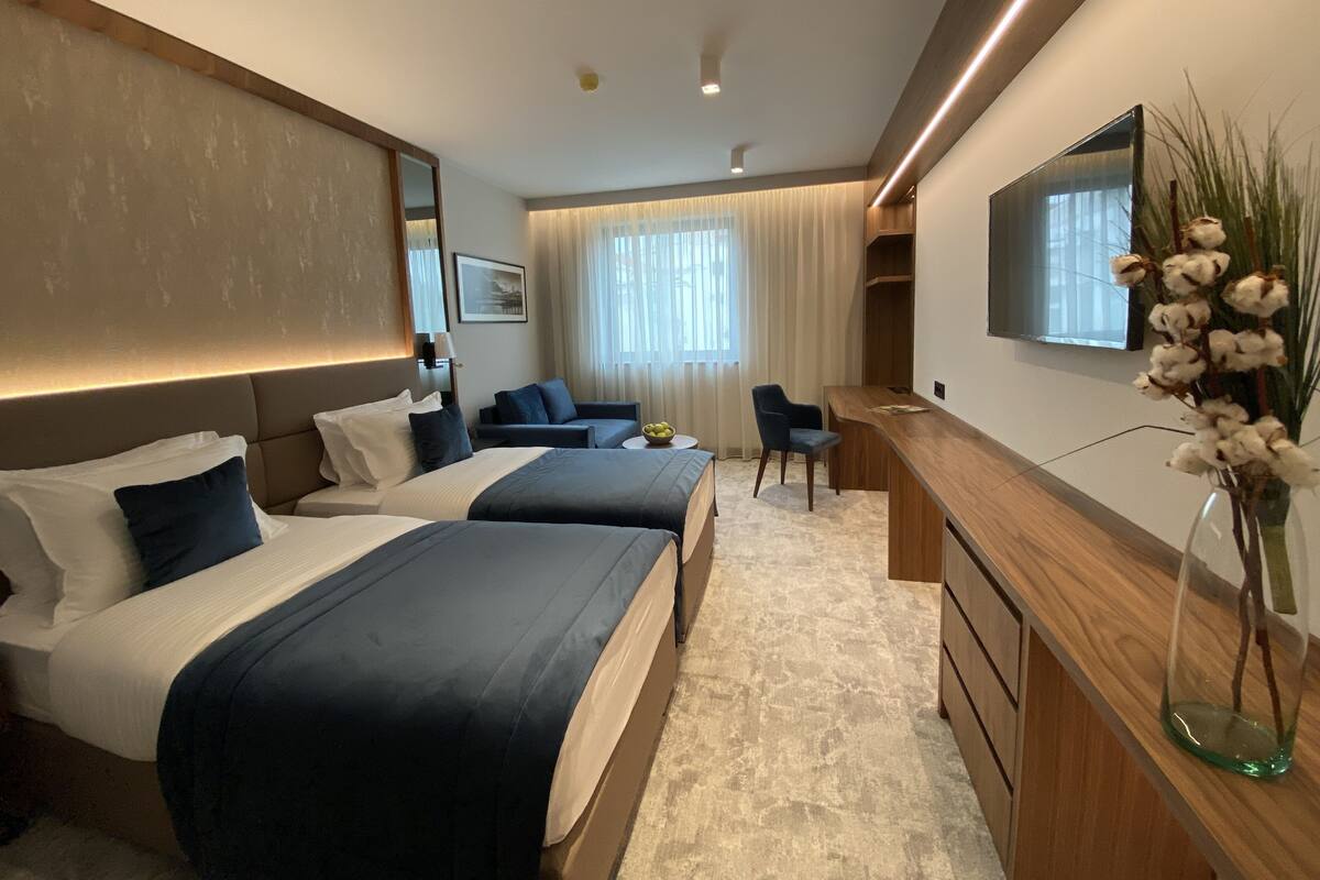 A modern hotel room with two beds, a desk, a chair, a TV, and a large window with curtains.