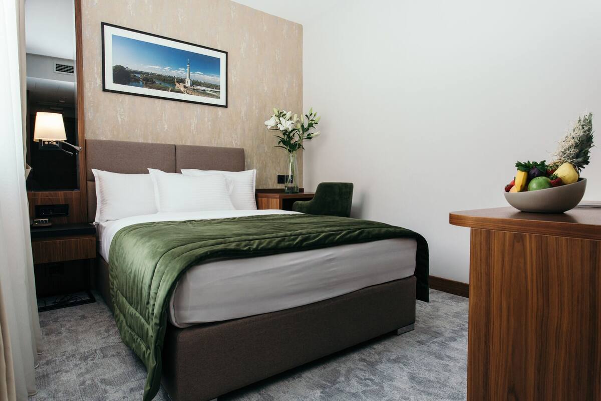 A hotel room with a bed, a nightstand with a lamp, a desk with a fruit bowl, and a framed picture on the wall.