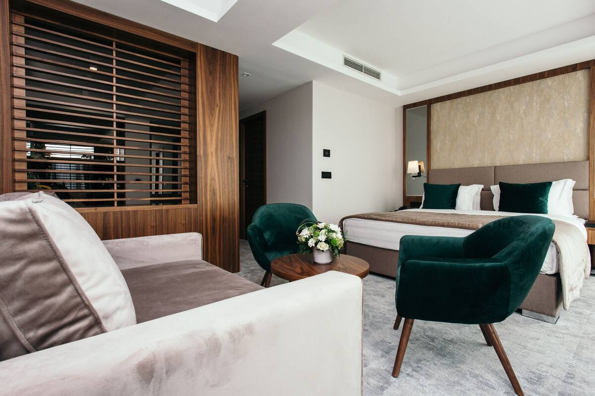 A modern hotel room with a large bed, a sofa, two armchairs, a small table with a vase of flowers, and a large window with blinds.