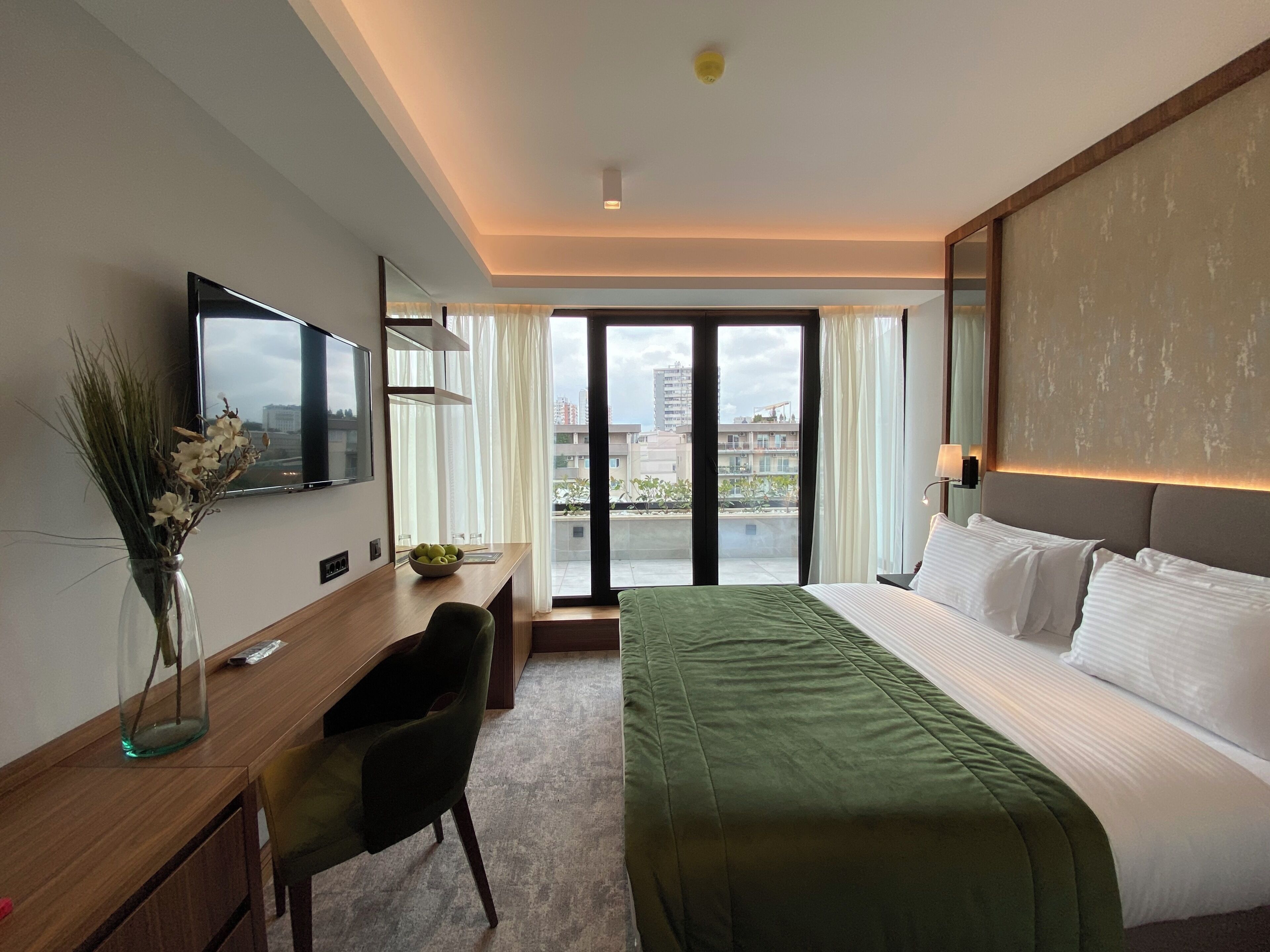 A hotel room with a bed, a desk, a chair, a TV, and a view of buildings through the window.