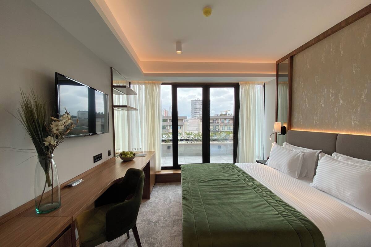 A hotel room with a bed, a desk, a chair, a TV, and a view of buildings through the window.