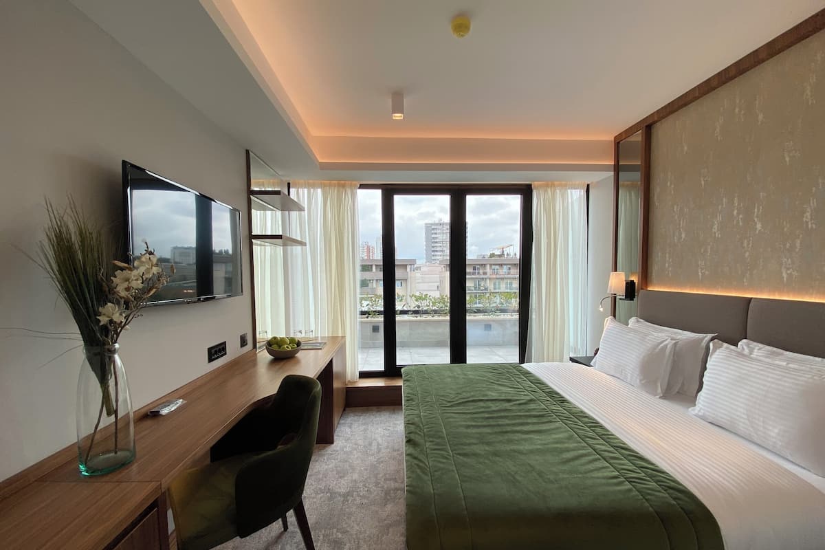 A hotel room with a bed, a desk, a chair, a TV, and a view of buildings through the window.
