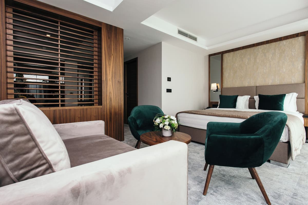 A modern hotel room with a bed, a sofa, a chair, a small table with a vase, and a large window with blinds.