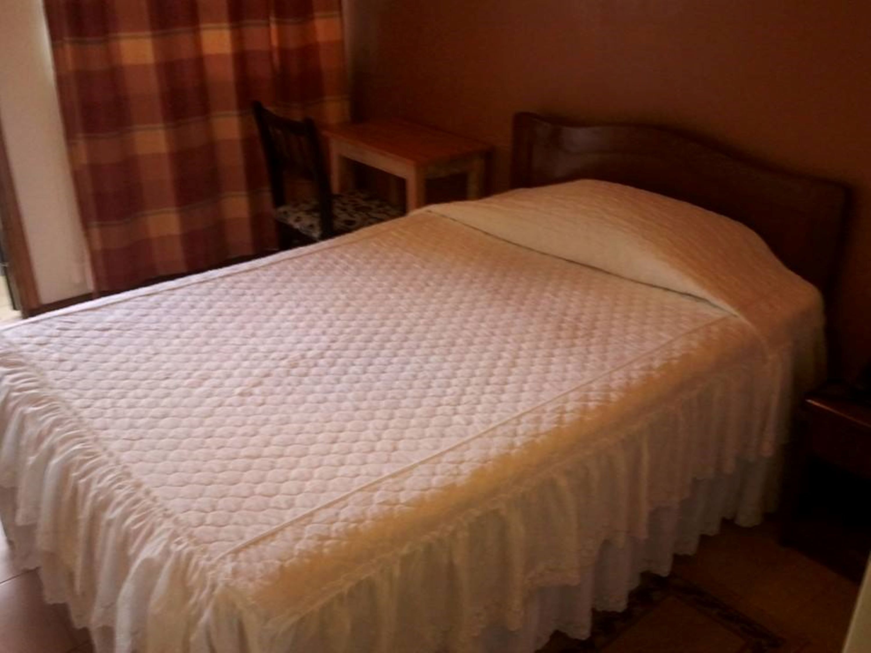 Una cama con cubrecolcha acolchada y bordes festoneados, una mesita de noche de madera y una silla con cojín estampado.