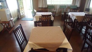 Un comedor con mesas y sillas de madera, un dispensador de agua y vistas a través de una ventana con cortinas.