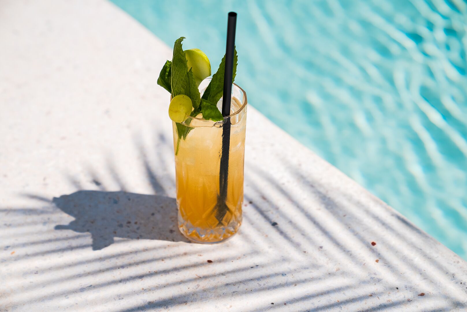 A glass of iced drink with mint leaves and a straw, placed on a white surface by a pool.