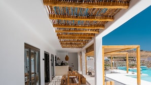 A modern outdoor patio with a wooden pergola, a pool, and a dining area with a view of the landscape. - Koukoumi Vegan Boutique Hotel (Mykonos)