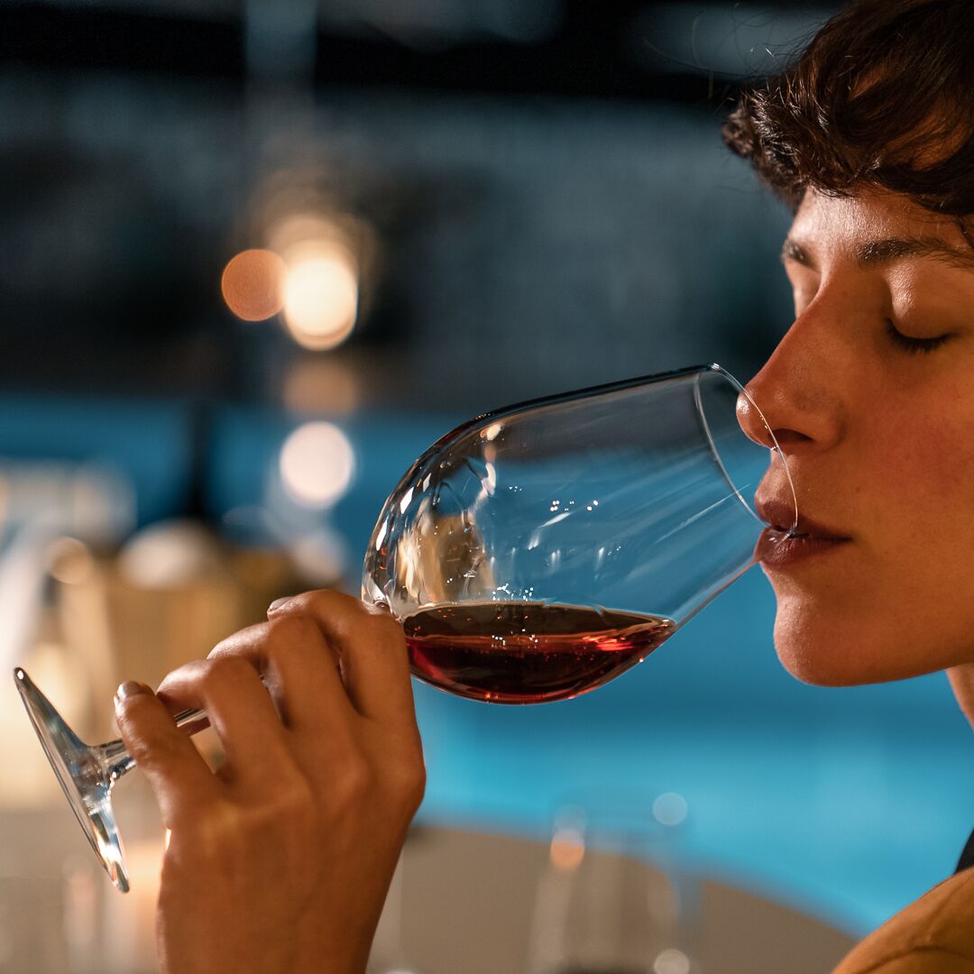Una persona sosteniendo una copa de vino con un líquido oscuro, probablemente vino, en un ambiente tenuemente iluminado, con una piscina al fondo.