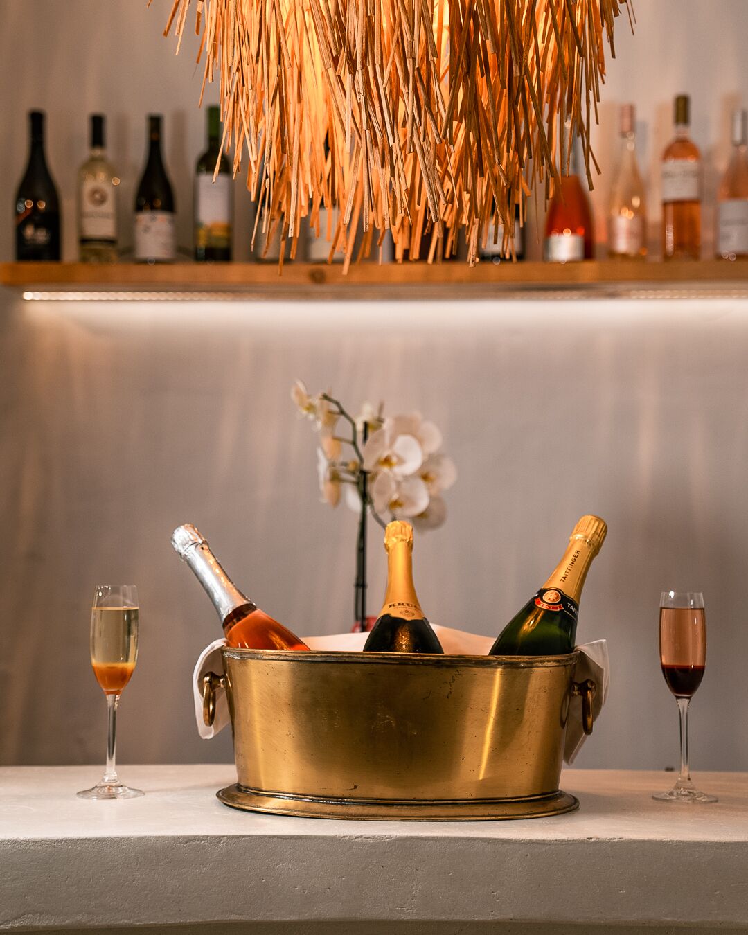 Un área de bar con un cubo metálico que contiene botellas de champán y copas, una estantería con varias botellas al fondo y una planta decorativa.