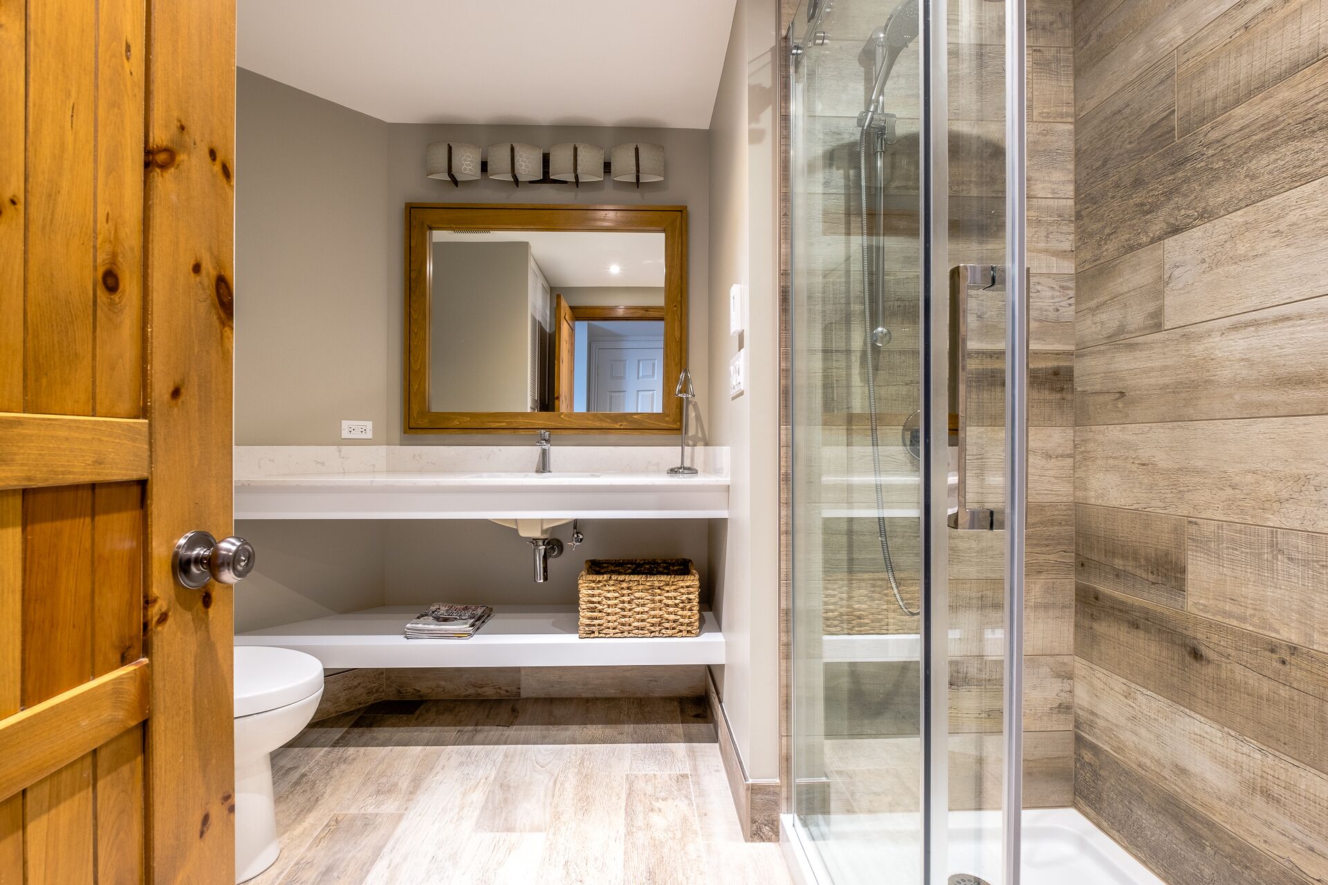 Une salle de bain moderne avec une porte en bois, un lavabo blanc, un miroir avec un cadre en bois, une cabine de douche en verre et un mur en bois.