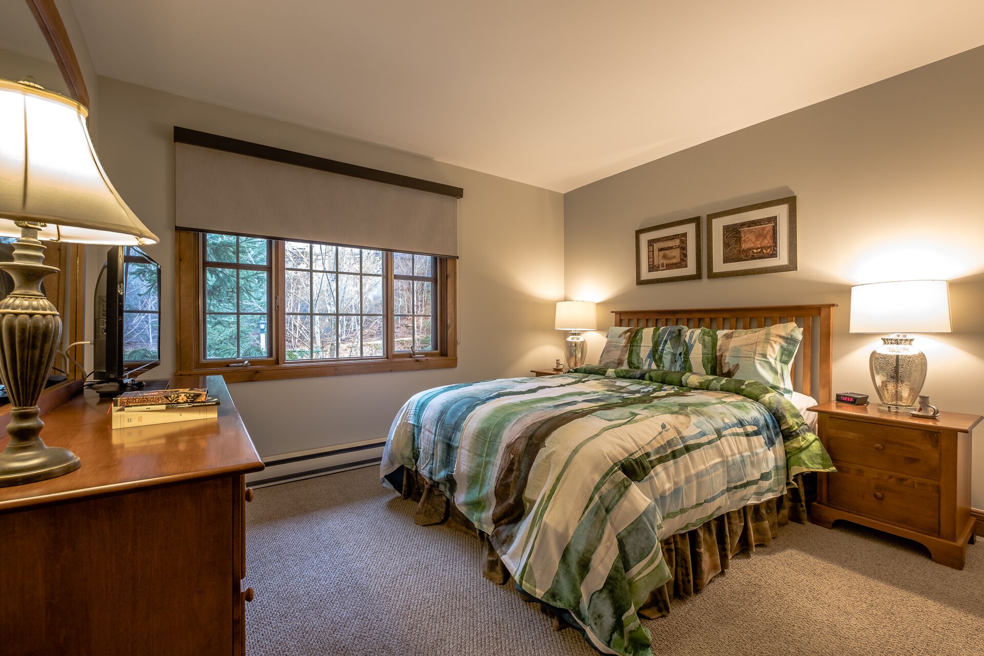A bedroom with a bed, two bedside lamps, a wooden nightstand, a desk with a lamp, and a window with a view of trees.